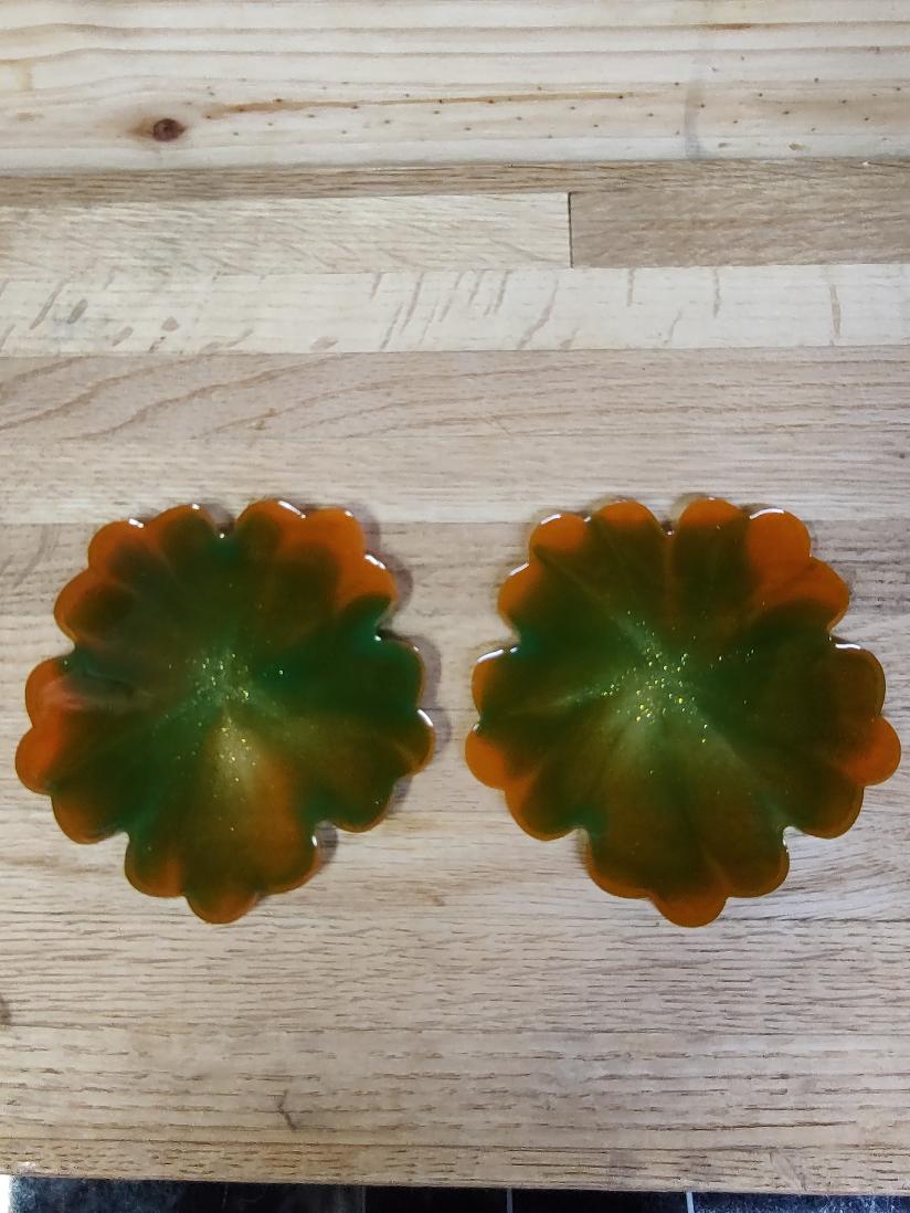 A pair of scalloped, flower-shaped glass coasters with a green and orange marbled pattern, displayed on a light wood grain surface.