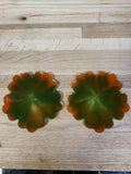 A pair of scalloped, flower-shaped glass coasters with a green and orange marbled pattern, displayed on a light wood grain surface.